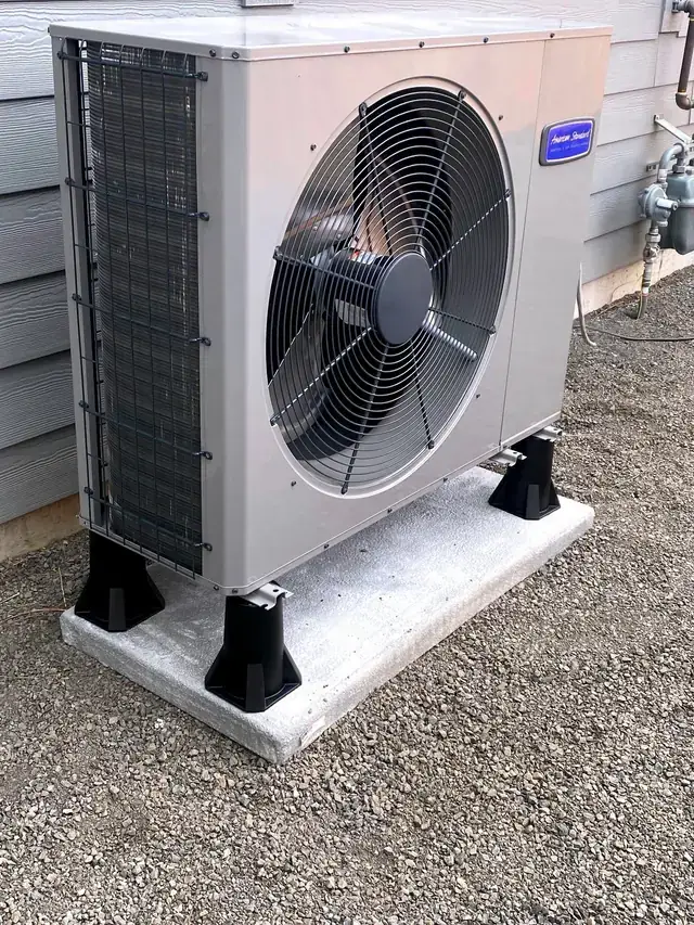 An American Standard heat pump outdoor unit set on a raised pad over gravel