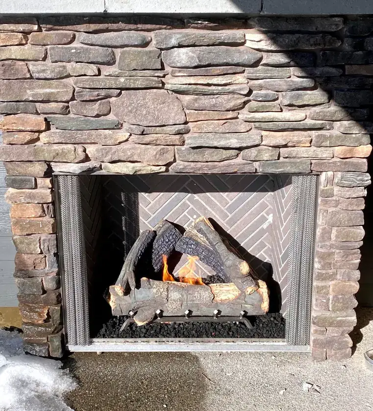 A lit outdoor gas fireplace in a stacked-stone surround with snow at its base